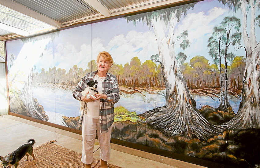 A TOUCH OF ART… Local resident, Joyce Yates holding her puppy, Theo in front of a giant mural of the Murray River she painted on a wall at her home in the Big 4 Shepparton Park Lane Holiday Park. Photo: Katelyn Morse.